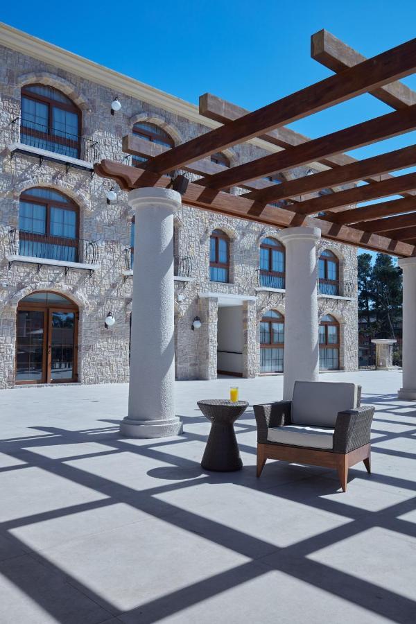 Buona Vitta Gramado Resort & Spa By Gramado Parks Exterior foto The photo shows a well-designed outdoor space with a stone wall building in the background. There are several tall columns that support a wooden pergola overhead, creating a shaded area. In this space, there is a stylish chair and a small table besid