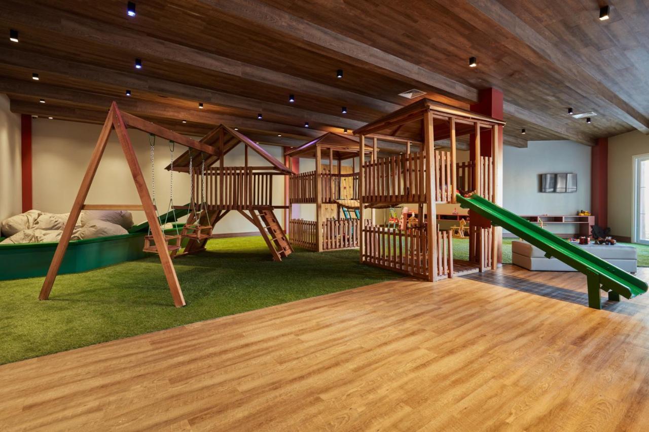 Buona Vitta Gramado Resort & Spa By Gramado Parks Exterior foto The photo shows an indoor play area designed for children. It features a wooden play structure that includes multiple levels, slides, and a swing set. The floor is covered with wooden planks, and there is a green carpet-like area that adds a soft tou