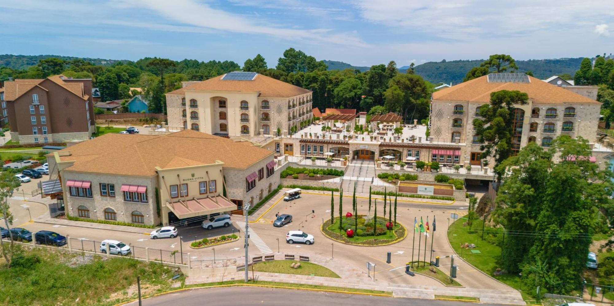 Buona Vitta Gramado Resort & Spa By Gramado Parks Exterior foto The photo shows an aerial view of a hotel complex surrounded by lush greenery. The buildings are designed with beige and light-colored facades, featuring multiple stories and a spacious layout. In the foreground, there is a circular road with several