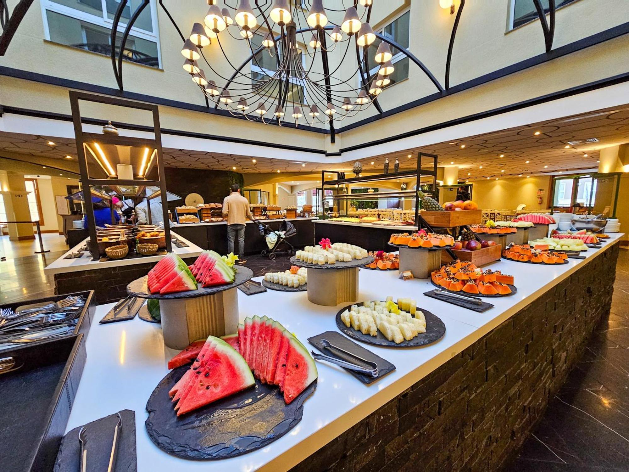 Buona Vitta Gramado Resort & Spa By Gramado Parks Exterior foto The photo depicts a lavish buffet setup in a dining area. In the foreground, there are several platters featuring an assortment of colorful food items, primarily fruits like watermelon and various types of sushi or rolls. The buffet table is elegantl