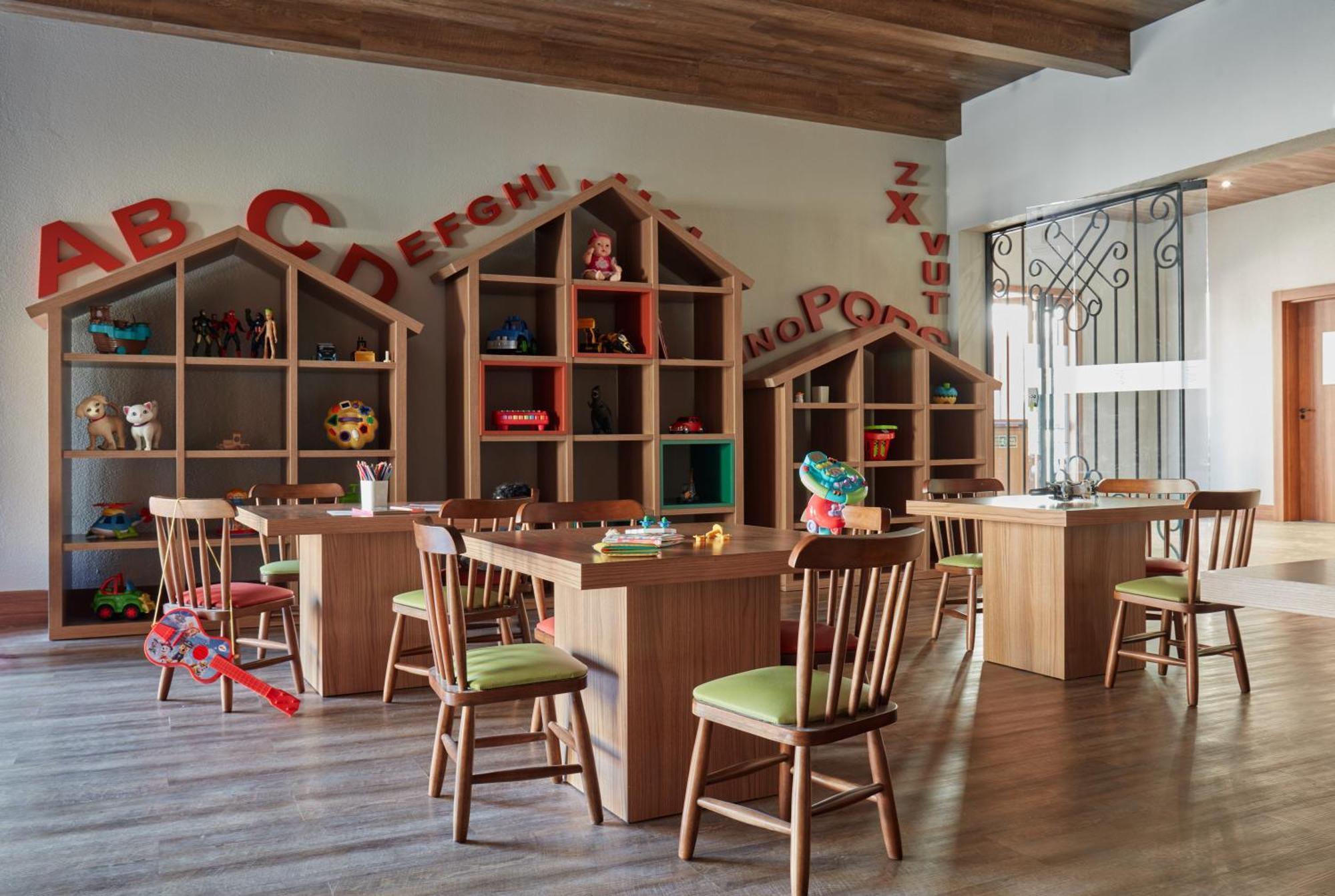 Buona Vitta Gramado Resort & Spa By Gramado Parks Exterior foto The photo shows a classroom or playroom designed for children. It features wooden furniture, including several tables and chairs with green cushions. In the background, there are large, open shelving units shaped like houses, displaying various toys 