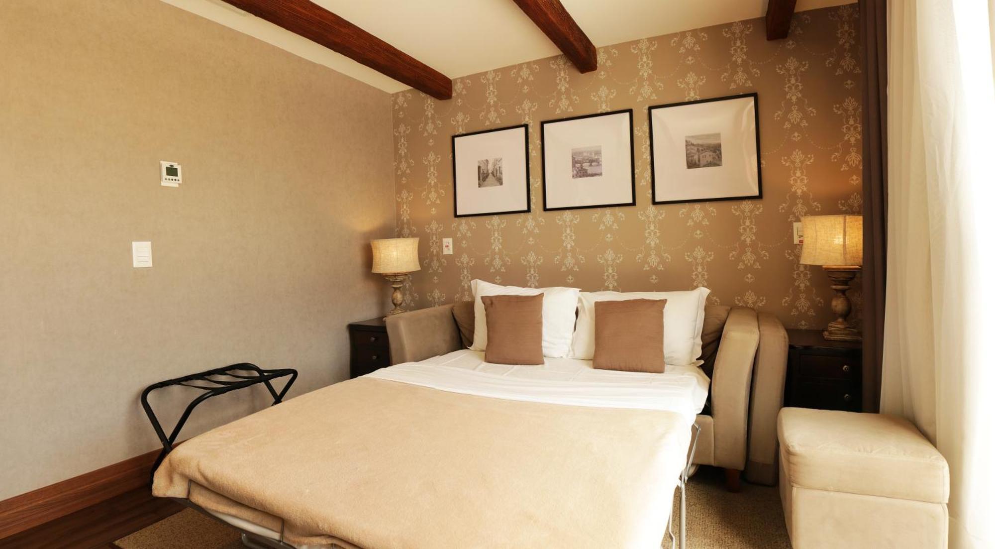 Buona Vitta Gramado Resort & Spa By Gramado Parks Exterior foto The photo shows a cozy bedroom with a neutral color palette. There is a bed with white linens and two brown decorative pillows. On either side of the bed, there are bedside tables with lamps. The walls feature a subtle floral pattern, and three frame