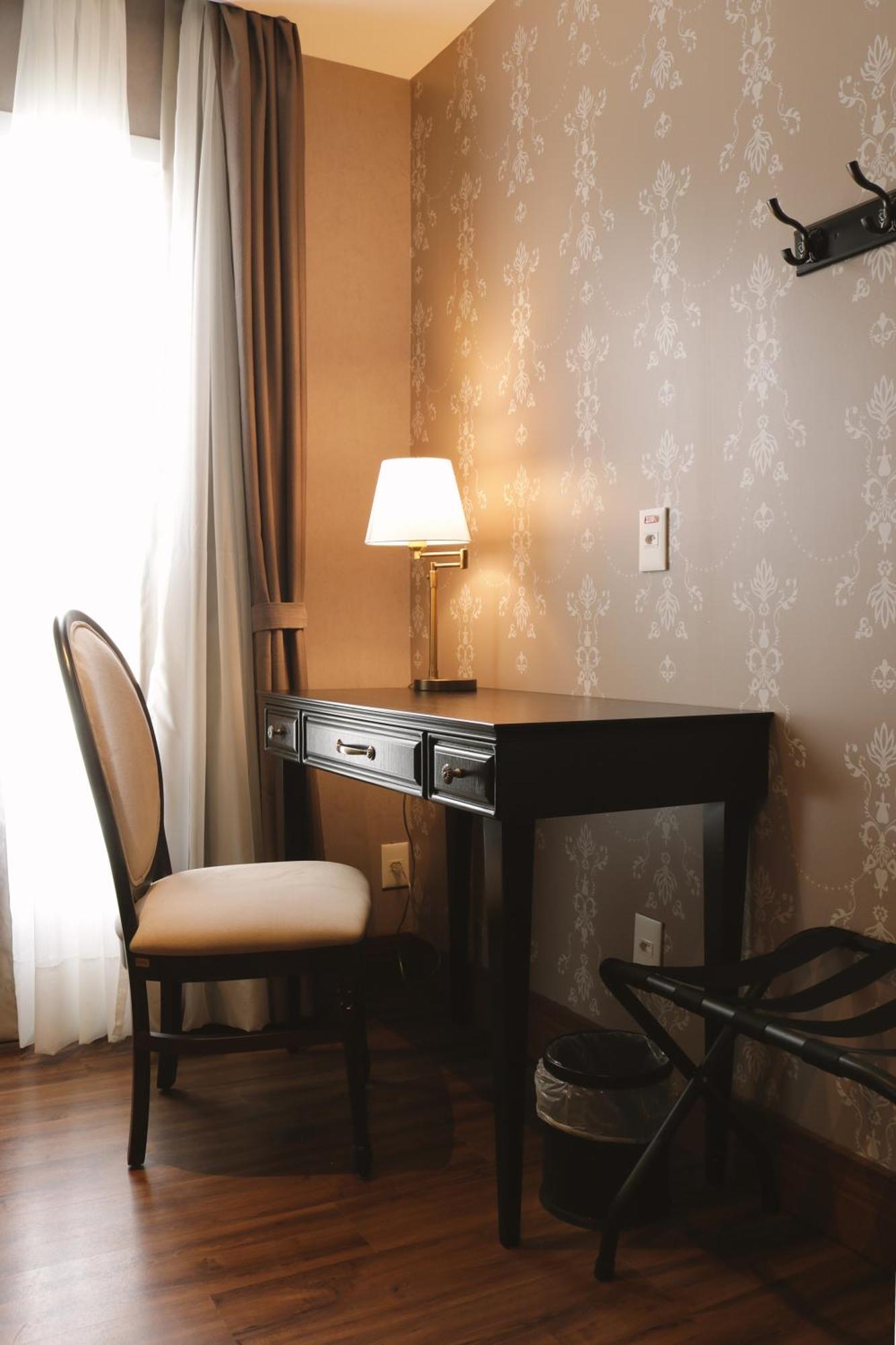 Buona Vitta Gramado Resort & Spa By Gramado Parks Exterior foto The photo shows a small desk set against a wall with a decorative pattern. The desk is black with a few drawers and has a small lamp on it. Next to the desk, there is a chair with a light-colored cushion. Soft light is coming through a window, castin