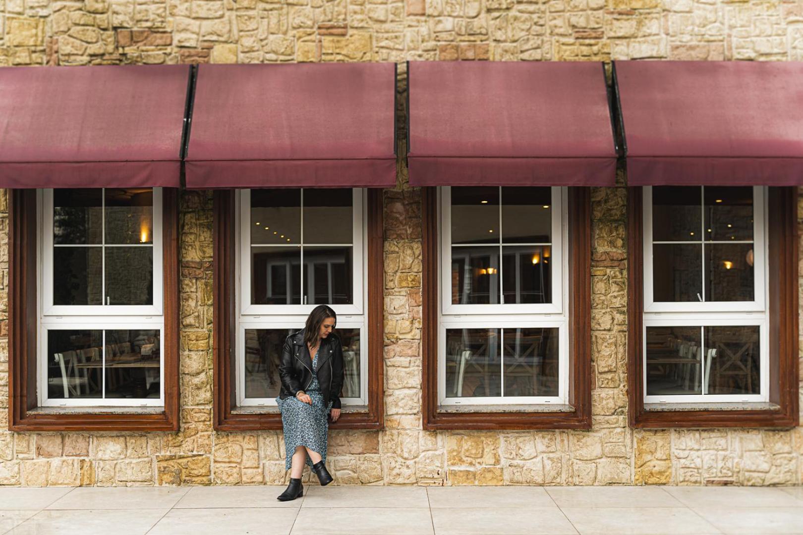 Buona Vitta Gramado Resort & Spa By Gramado Parks Exterior foto The photo shows a woman standing against a stone wall with three large windows in front of her, each framed with wooden borders. The windows have maroon awnings above them. The woman is dressed in a long skirt and leather jacket, leaning slightly aga